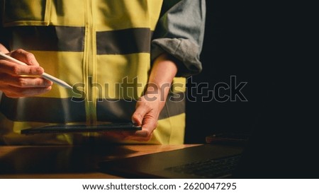 Construction worker in safety vest using digital pen and tablet at desk beside laptop under warm lighting for safety planning concept