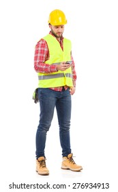 Construction Worker Sending A Text Message. Manual Worker In Yellow Helmet And Lime Waistcoat Using A Smart Phone. Full Length Studio Shot Isolated On White.