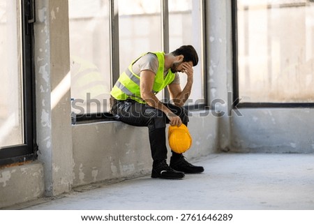 A construction worker sits on a ledge, holding a yellow hard hat. He appears tired and takes a break from work inside a building under construction. The walls are bare and bright.