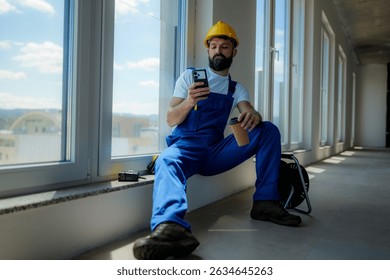 Construction worker sitting by a window, savoring a coffee break while using a smartphone in a modern building under construction. Enjoying a moment of relaxation amidst the busy site