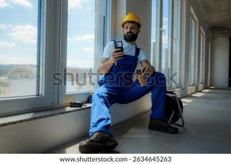 Construction worker sitting by a window, savoring a coffee break while using a smartphone in a modern building under construction. Enjoying a moment of relaxation amidst the busy site
