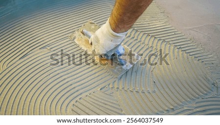 Construction worker spreading tile adhesive on floor using notched trowel, preparing surface for tiling