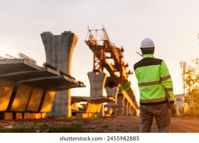 A construction worker is standing on a bridge, talking on his cell phone. The scene is set in a construction site, with a large crane in the background. The worker is wearing a yellow jacket