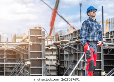 A construction worker stands confidently at a building site, observing the progress of the structure. Dressed in safety gear including a helmet and gloves, he watches as machinery operates