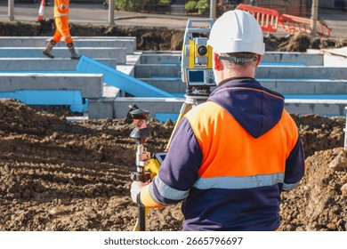 Construction worker stands focused on surveying equipment at a construction site. The worker wears safety gear and oversees groundwork for a new building