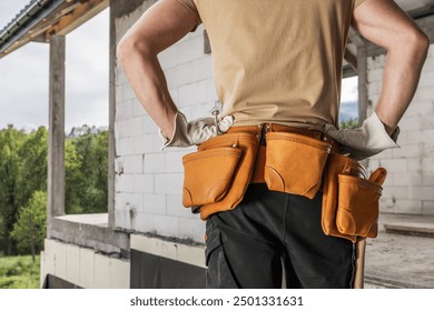 A construction worker stands with hands on hips, wearing a tool belt filled with equipment at a building site. - Powered by Shutterstock