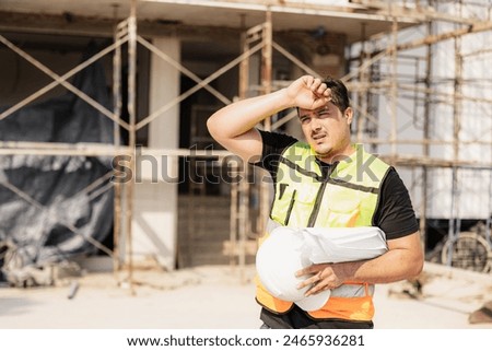Construction worker taking a break and sweating under the sun, holding a hard hat, with construction scaffolding in the background.