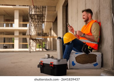 Construction worker taking a break from work, sitting and enjoying a cup of coffee while checking messages on his smartphone, with his toolbox and hardhat nearby