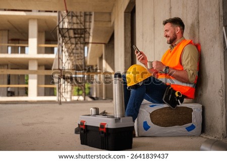 Construction worker taking a break from work, sitting and enjoying a cup of coffee while checking messages on his smartphone, with his toolbox and hardhat nearby