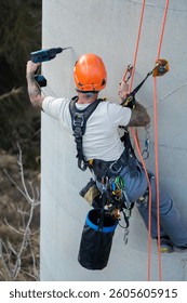 Construction worker with tattoos using a drill while hanging on ropes and wearing safety equipment