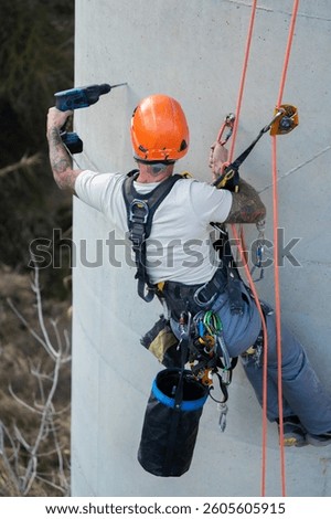 Construction worker with tattoos using a drill while hanging on ropes and wearing safety equipment