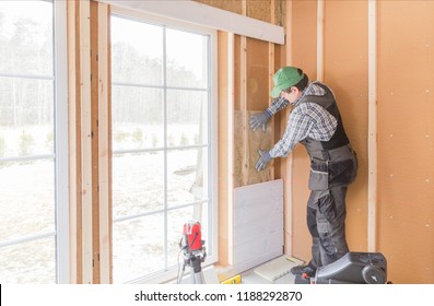 Construction worker thermally insulating eco-wood frame house with wood fiber plates and heat-isolating natural hemp material. Finishing the walls with a white wooden board, using laser line level.