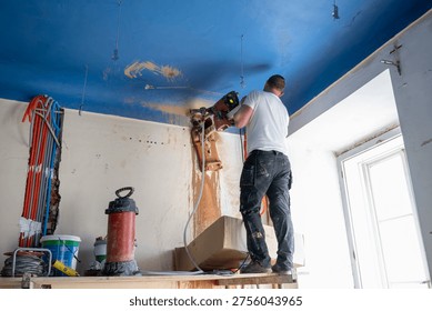 Construction worker uses a core drilling rig to cut a ceiling penetration for future mechanical routing, while slurry and dust mark the active work zone near the window.