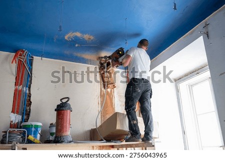 Construction worker uses a core drilling rig to cut a ceiling penetration for future mechanical routing, while slurry and dust mark the active work zone near the window.