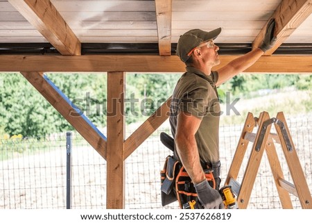 A construction worker uses a tool to secure wooden panels while standing on a ladder in a rural setting during the day. The environment is bright and inviting.