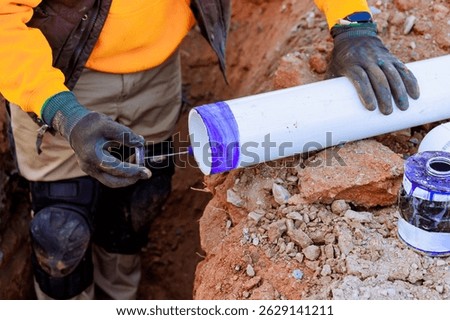 Construction worker is using blue adhesive on white PVC pipe on set up drainage pipe system in trench