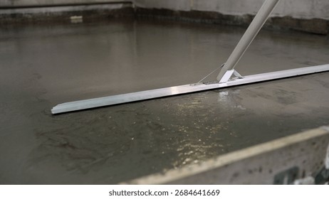 Construction worker is using a bull float to level a freshly poured concrete slab.
