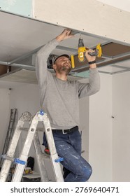 Construction worker using drill on metal profile frame for drywall suspended ceiling system near air duct and gypsum board panels. Installation process in an apartment under renovation
