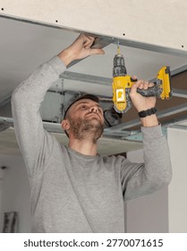 Construction worker using drill on metal profile frame for drywall suspended ceiling system near air duct and gypsum board panels. Installation process in an apartment under renovation