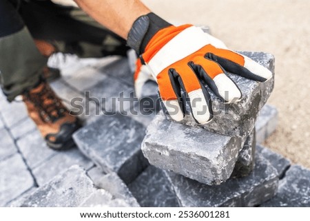 A construction worker using gloved hands carefully positions gray paving stones on a worksite, showcasing hard labor and attention to detail during the daytime.