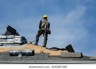 Construction worker using pneumatic nailer and safety helmet applying roofing materials against blue sky backdrop