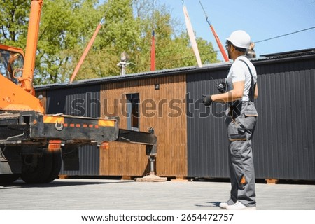 Construction worker using remote control, managing crane lifting prefabricated modular house onto truck, showcasing efficient building transport