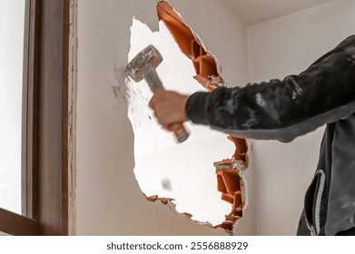 Construction worker using sledgehammer to demolish interior brick wall, creating hole during home renovation