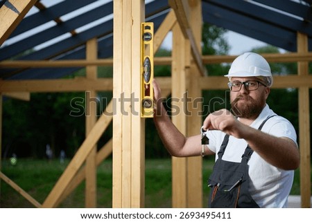 Construction worker using spirit level to check wooden beam at building site while holding keys. Builder in safety helmet and overalls working on timber frame house construction outdoors.