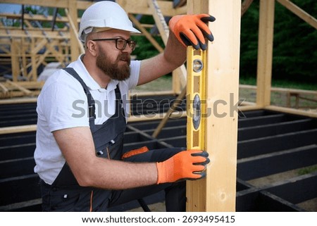 Construction worker using a spirit level to check vertical alignment of wooden beams at a building site. Man wearing protective helmet and glasses working on wooden frame structure. 