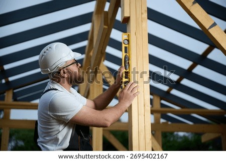 Construction worker using spirit level to check vertical alignment of wooden beam at frame house site. Builder wearing hard hat and safety glasses working on wooden structure outdoors.