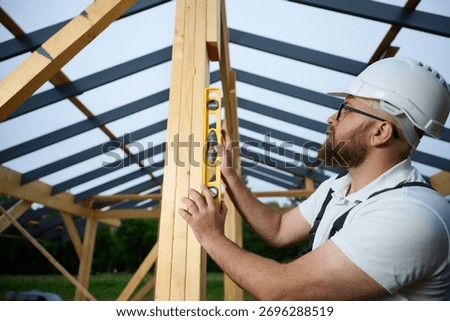 Construction worker using spirit level to check vertical alignment of wooden beam at frame house site. Builder wearing hard hat and safety glasses working on wooden structure outdoors.