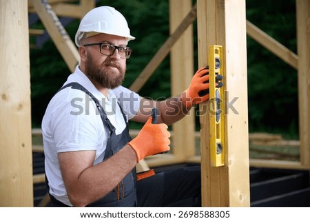 Construction worker using spirit level to check vertical alignment of wooden beam at frame house site. Builder wearing hard hat and safety glasses working on wooden structure outdoors.
