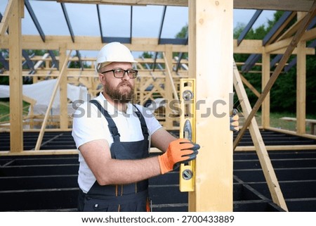 Construction worker using spirit level to check vertical alignment of wooden beam at frame house site. Builder wearing hard hat and safety glasses working on wooden structure outdoors.