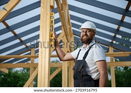 Construction worker using spirit level to check vertical alignment of wooden beam at frame house site. Builder wearing hard hat and safety glasses working on wooden structure outdoors.
