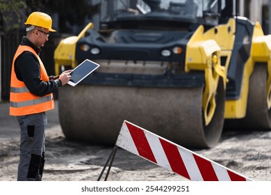 Construction worker using a tablet while overseeing roadwork with a steamroller in the background
