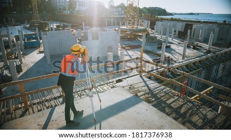 Construction Worker Using Theodolite Surveying Optical Instrument for Measuring Angles in Horizontal and Vertical Planes on Construction Site. Worker in Hard Hat Making Projections for the Building.