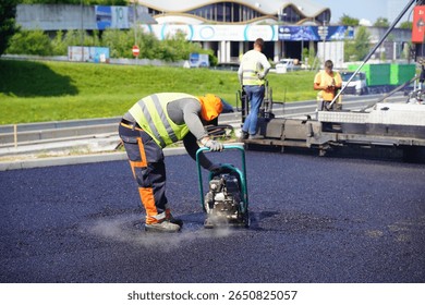 Construction Worker Using Vibratory Plate Compactor on Asphalt with Paver in the Background	