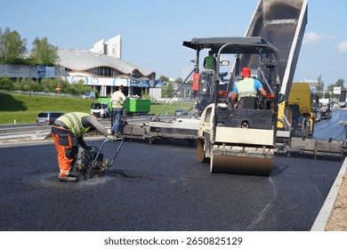 Construction Worker Using Vibratory Plate Compactor on Asphalt with Paver in the Background	