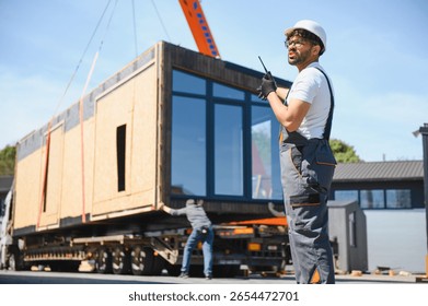 Construction worker using walkie talkie, coordinating loading of prefabricated house section onto truck, showcasing efficiency in modern building transport