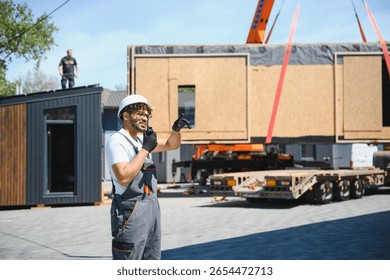Construction worker using walkie talkie and pointing while coordinating placement of prefabricated modular house section onto truck