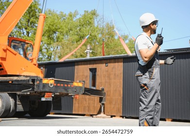 Construction worker using walkie talkie while supervising crane lifting prefabricated modular house onto truck in construction site