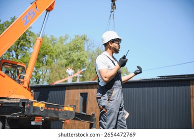 Construction worker using walkie talkie, coordinating crane lifting prefabricated house onto truck, showcasing teamwork and efficiency in modern housing construction