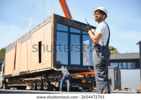 Construction worker using walkie talkie, coordinating loading of prefabricated house section onto truck, showcasing efficiency in modern building transport