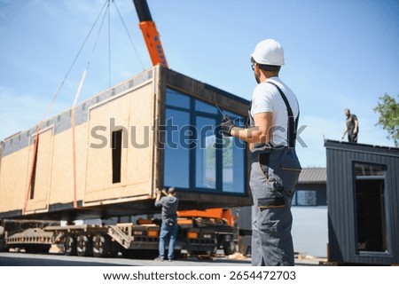 Construction worker using walkie talkie, supervising loading of prefabricated modular house onto truck in construction site