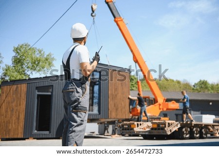 Construction worker using walkie talkie, coordinating crane lifting prefabricated modular house onto truck, showcasing efficient building transport