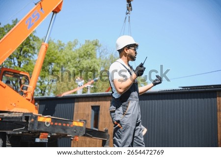 Construction worker using walkie talkie, coordinating crane lifting prefabricated house onto truck, showcasing teamwork and efficiency in modern housing construction