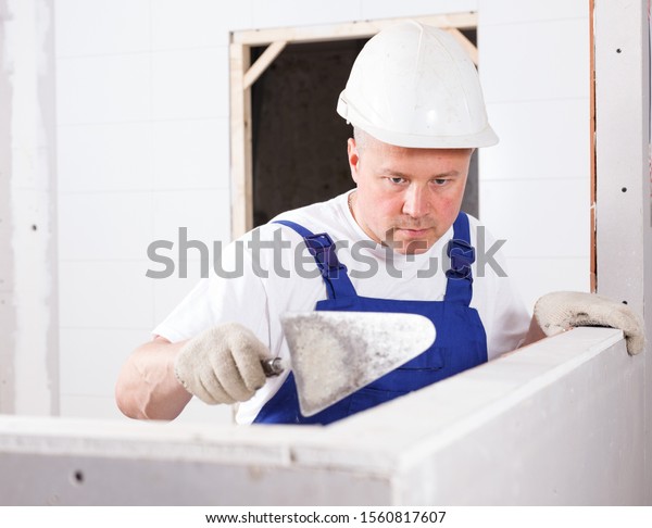 Construction worker with wall\
plastering tools mudding sheetrock wall in repairable\
room\
