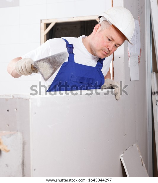 Construction worker with wall\
plastering tools mudding sheetrock wall in repairable\
room\
