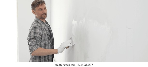 Construction worker wearing gloves and using a putty knife to plaster a white wall, performing home renovation and improvement tasks with precision