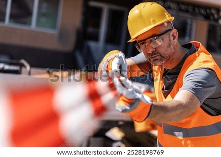 A construction worker wearing a hard hat, safety glasses, and a bright orange vest pulls on a bright orange safety barrier.